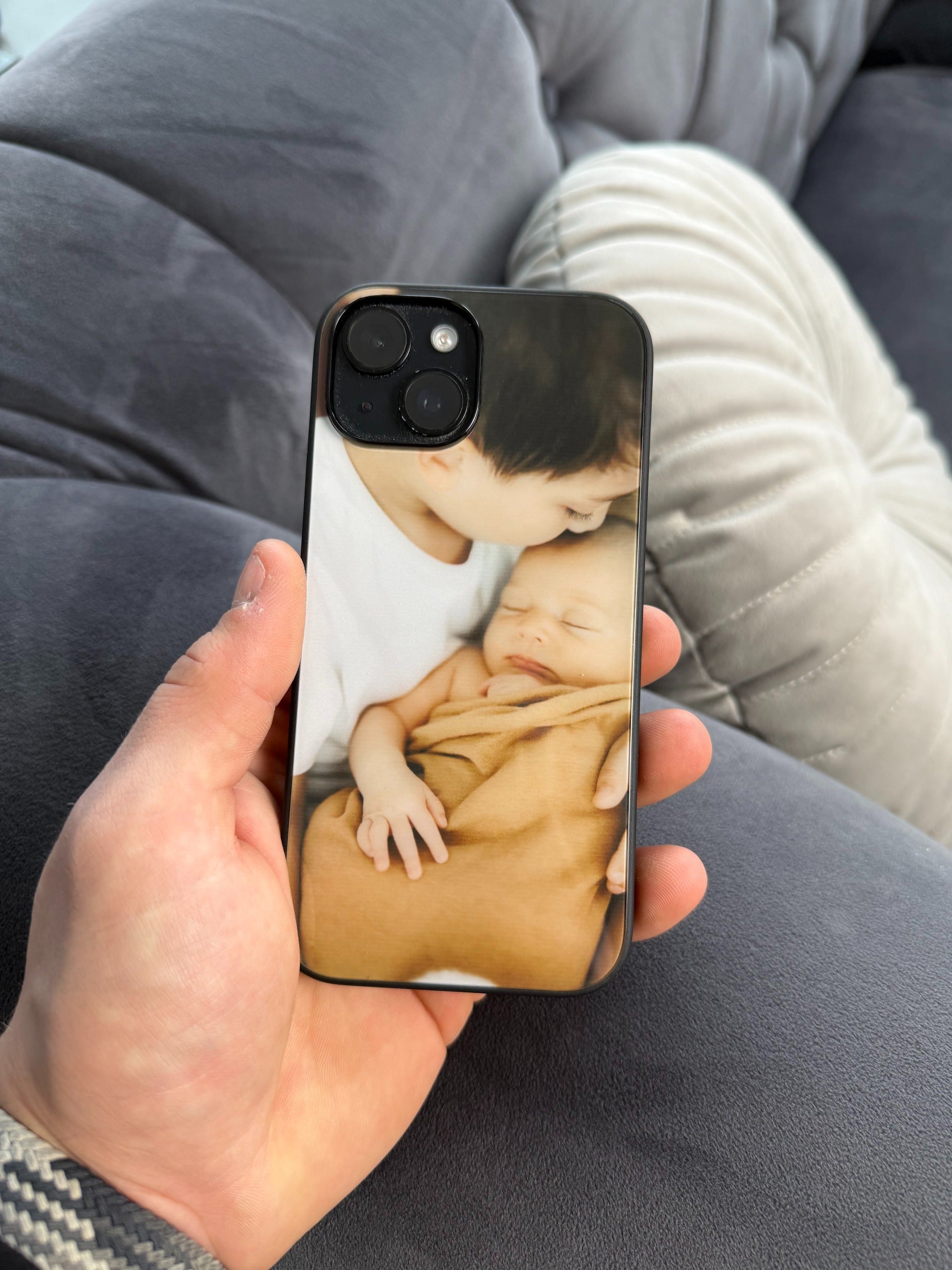 Hand hält eine personalisierte GREENMNKY Handyhülle mit Baby Foto Motiv, Nahaufnahme der Hülle mit Zweifachkamera Ausschnitt auf Sofa Hintergrund
