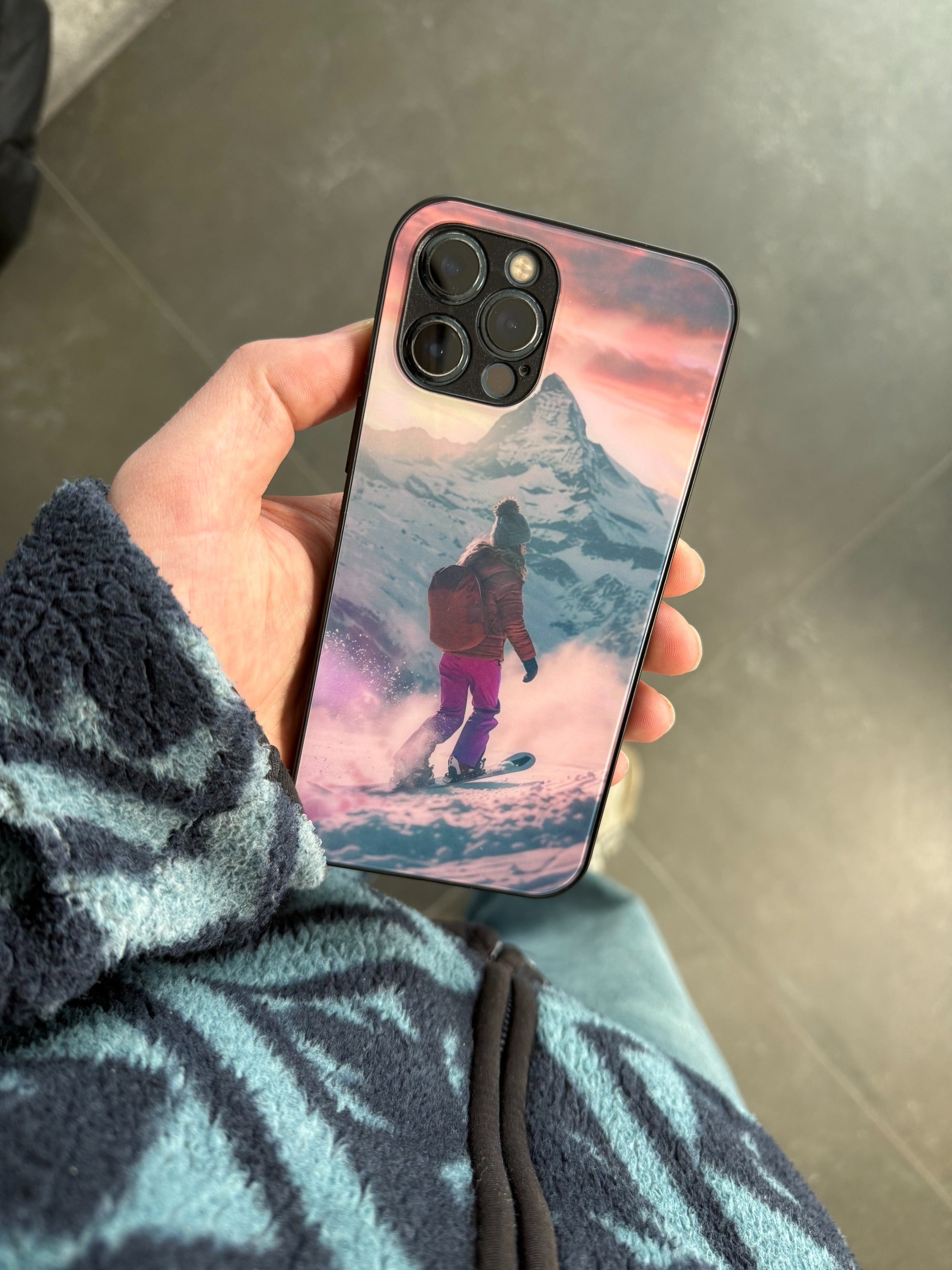 Hand hält eine personalisierte GREENMNKY Handyhülle mit Winterberg und Snowboard Motiv, Dreifachkamera Ausschnitt, Nahaufnahme auf grauem Boden