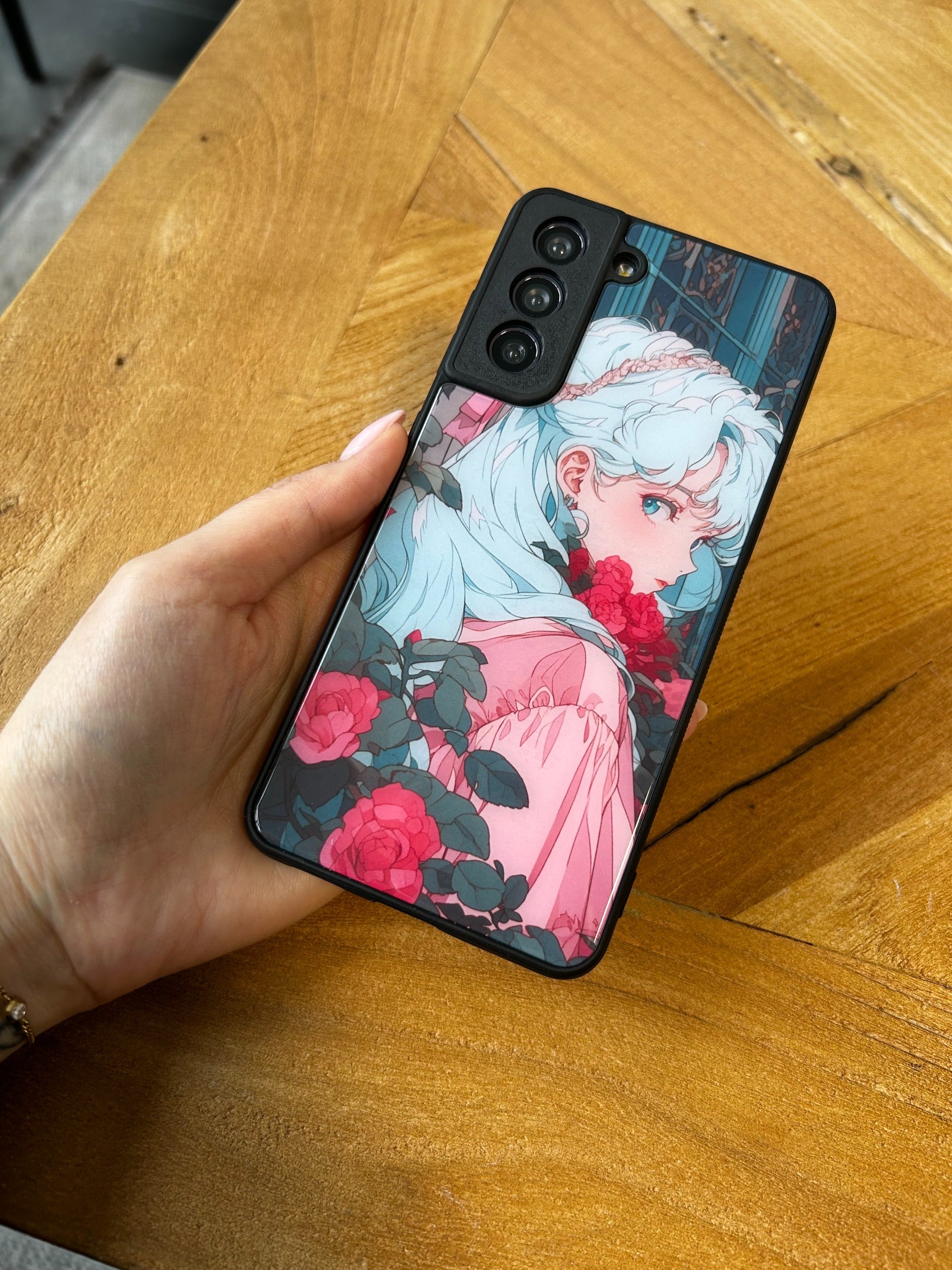 Hand hält eine personalisierte GREENMNKY Samsung Handyhülle mit Anime Illustration Motiv und rosa Blumen, Mehrfachkamera Ausschnitt sichtbar auf Holztisch Hintergrund